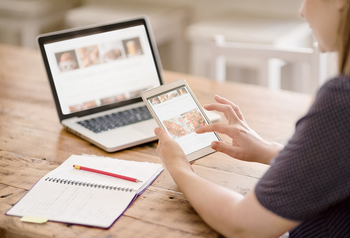 Cropped shot of the rear view of a female blogger reading information from social media holding digital tablet, with book and laptop in table.